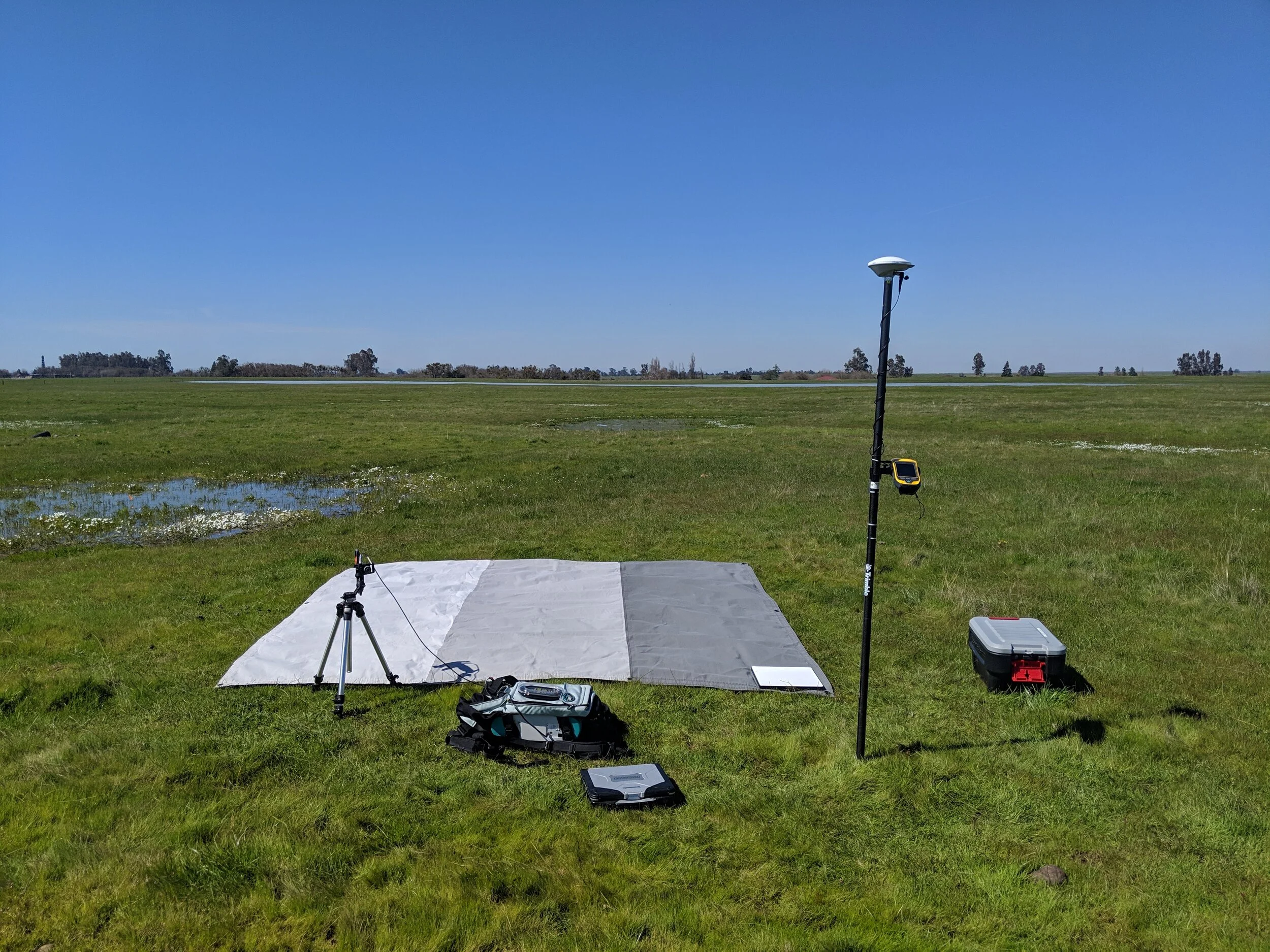 Hyperspectral field spectrophotometer, calibration panel, and GPS pole set up in a wet field